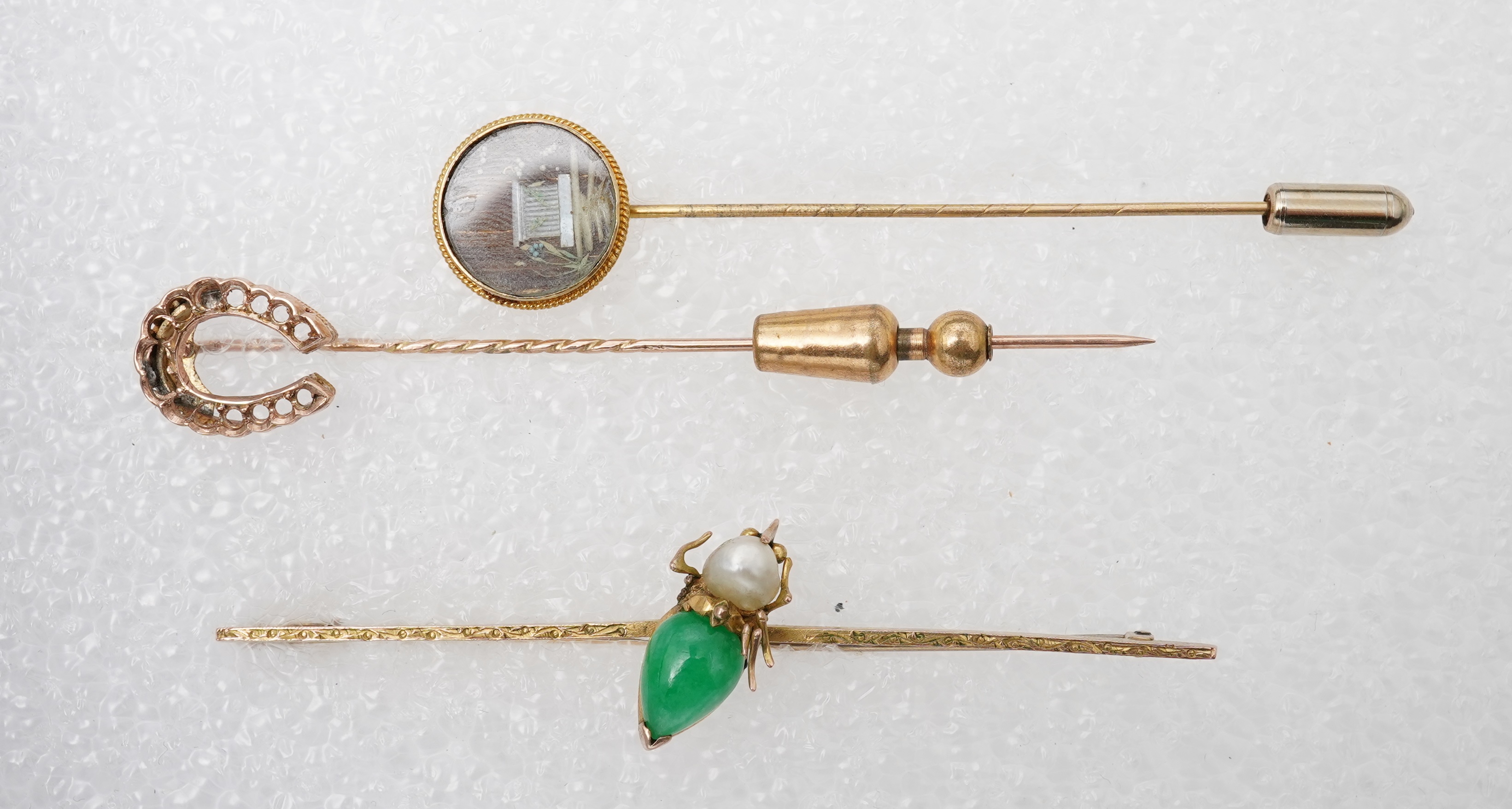 Two stick pins and a jadeite bar brooch, comprising: a bar brooch centring on an insect set with a pear-shape jadeite cabochon and a pearl, mounted in gold, length 7.5cm, stamped 14K, base metal pin; a stick pin capped w